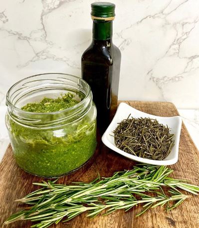 Clear glass jar of green pesto, fresh rosemary, dried rosemary and a bottle of olive oil on a wooden board