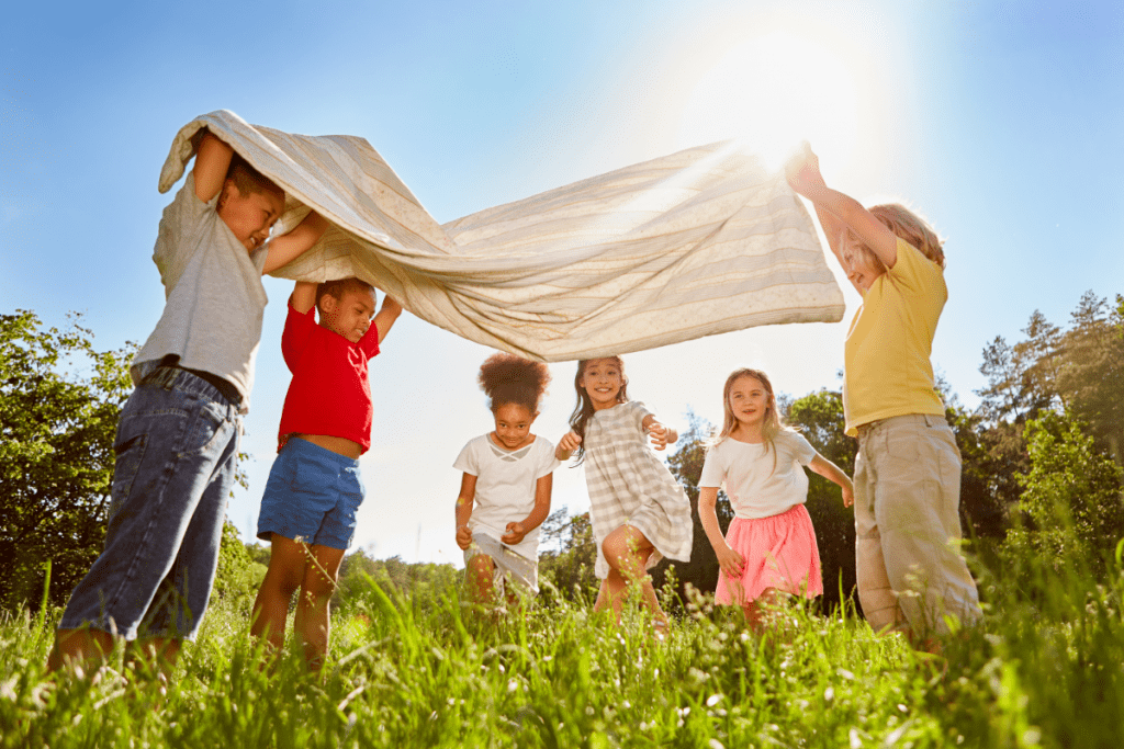 Teaching Kids About Sustainability - Group of happy kids playing in a field with a towel held up like a tent over them