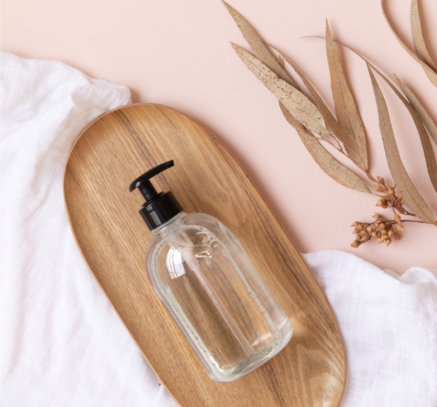 Natural Hand Sanitizers - Stock Image from Canva of a clear bottle of sanitizer on a wood board with a pink background, with a while piece of fabric and dried leaves
