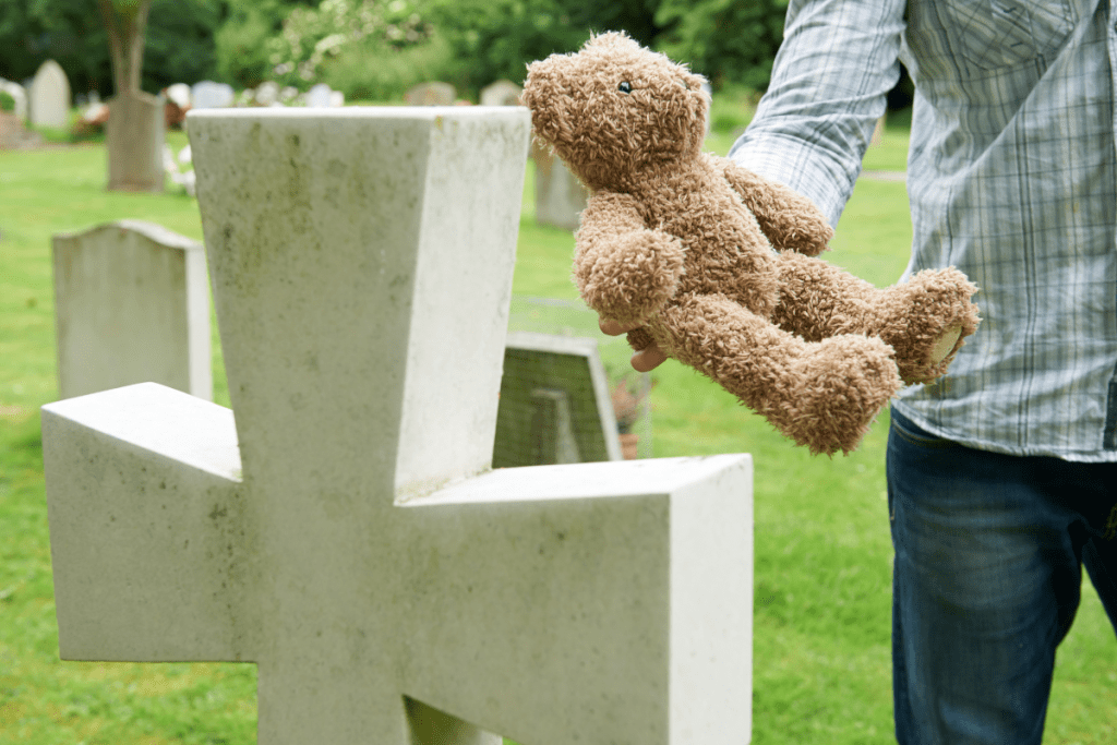 Image of a cross shaped gravestone and a man's arm holding out a brown teddybear to place in front of it