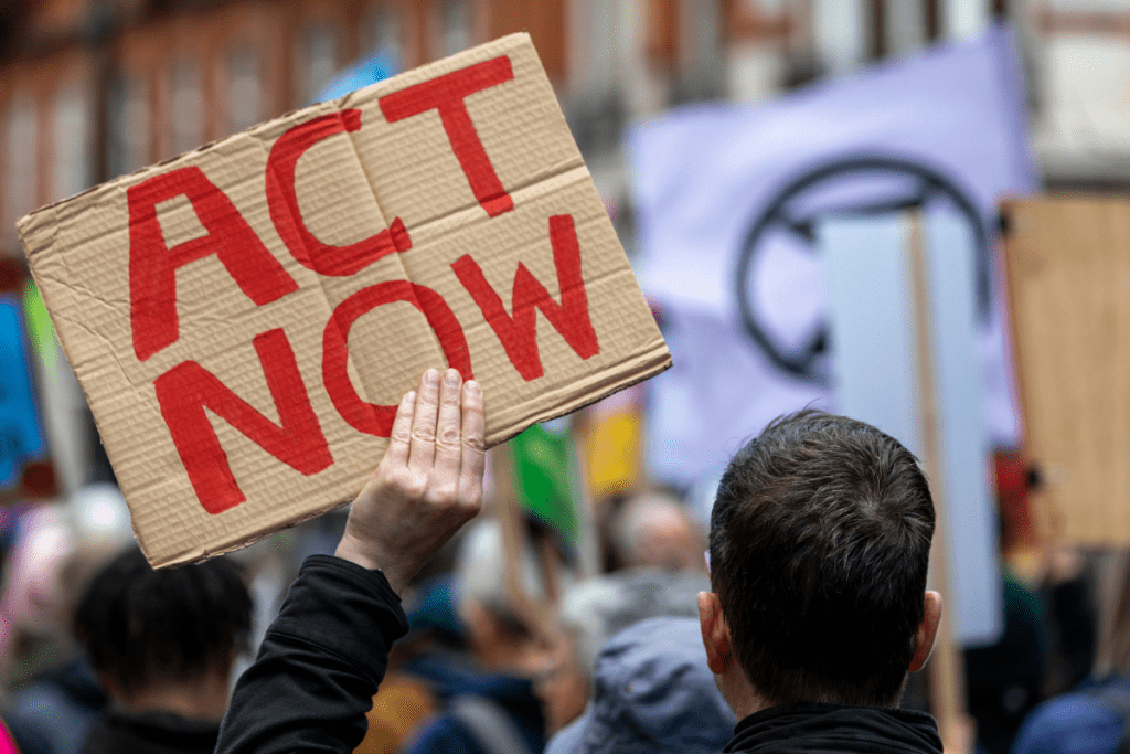 Cardboard placard held overhead during a climate march that says "Act Now" in red